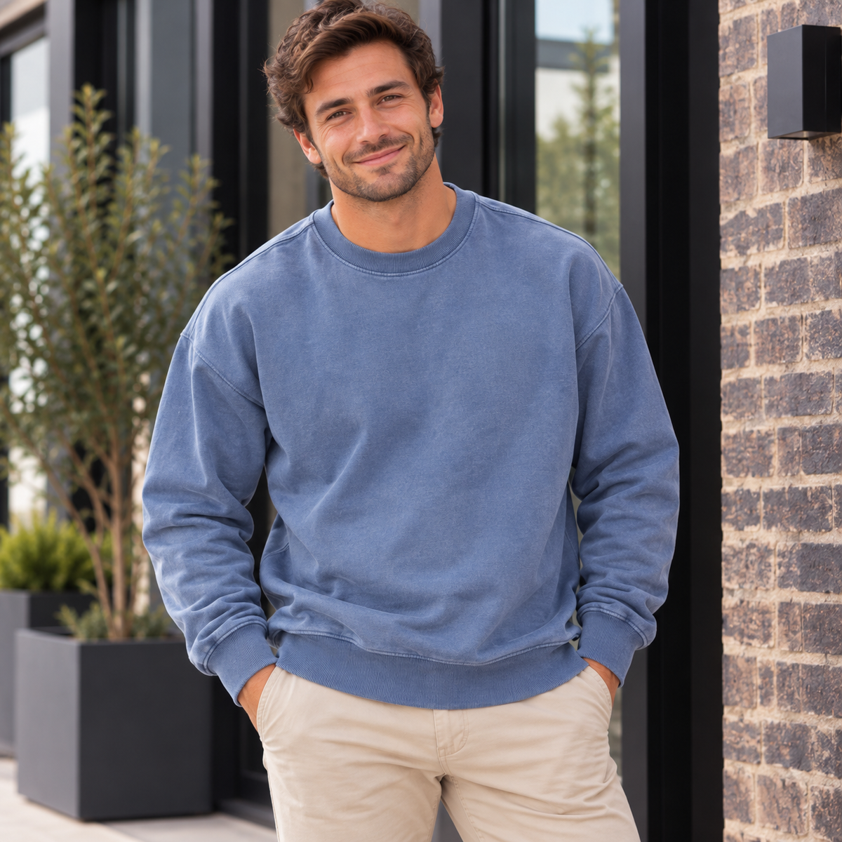 A man wearing a dusty blue crewneck sweatshirt and beige pants, standing outdoors in front of a modern building with a brick wall and potted plants.