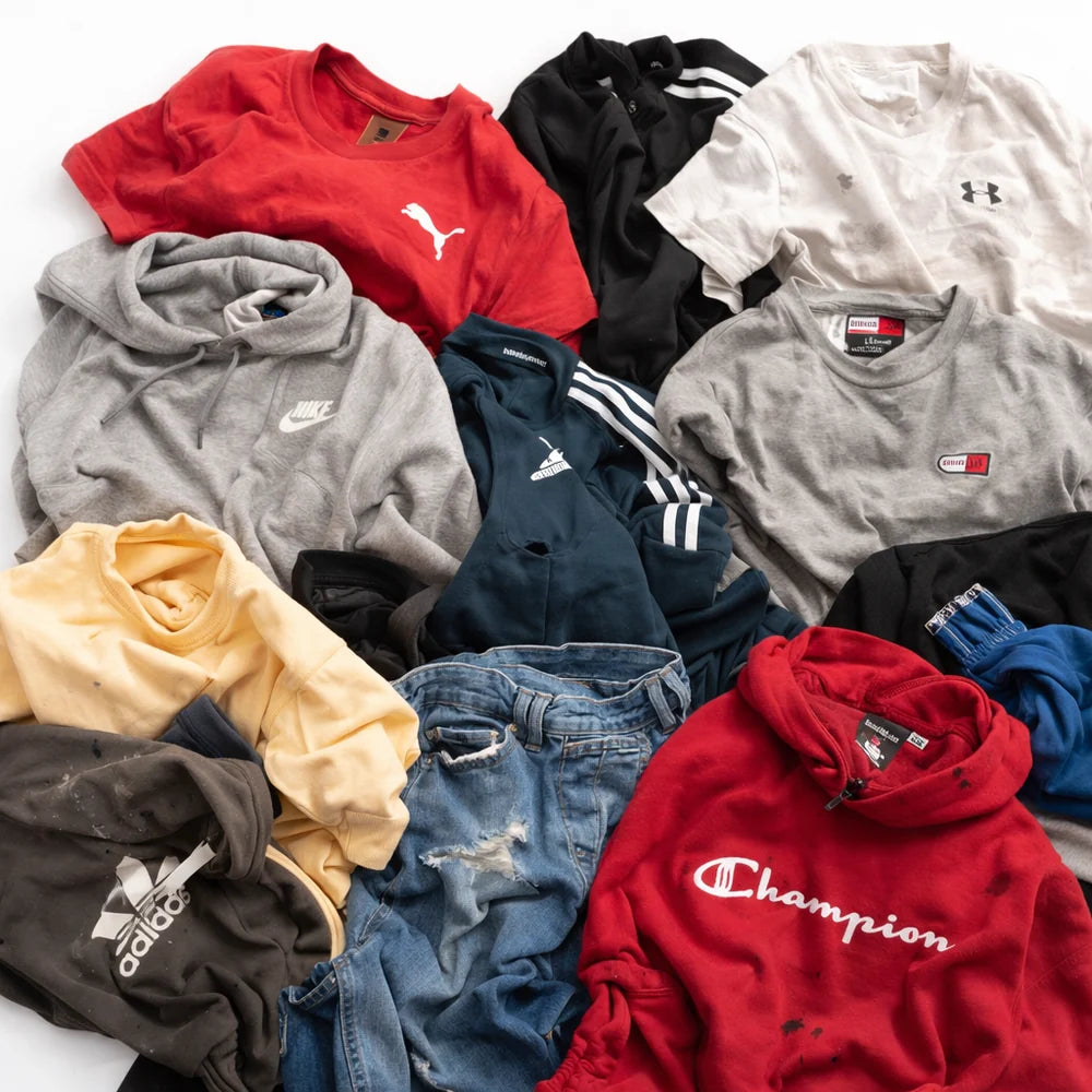 A pile of assorted vintage athletic wear and casual clothing, including a red Puma shirt, a gray Nike hoodie, a black Adidas track jacket, a gray Tommy Hilfiger shirt, a Champion hoodie, and distressed blue jeans, arranged on a white background.