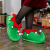A person wearing green elf slippers with red trim and white pom-poms, paired with black glitter tights, standing on a wooden floor with Christmas decorations and wrapped gifts in the background.