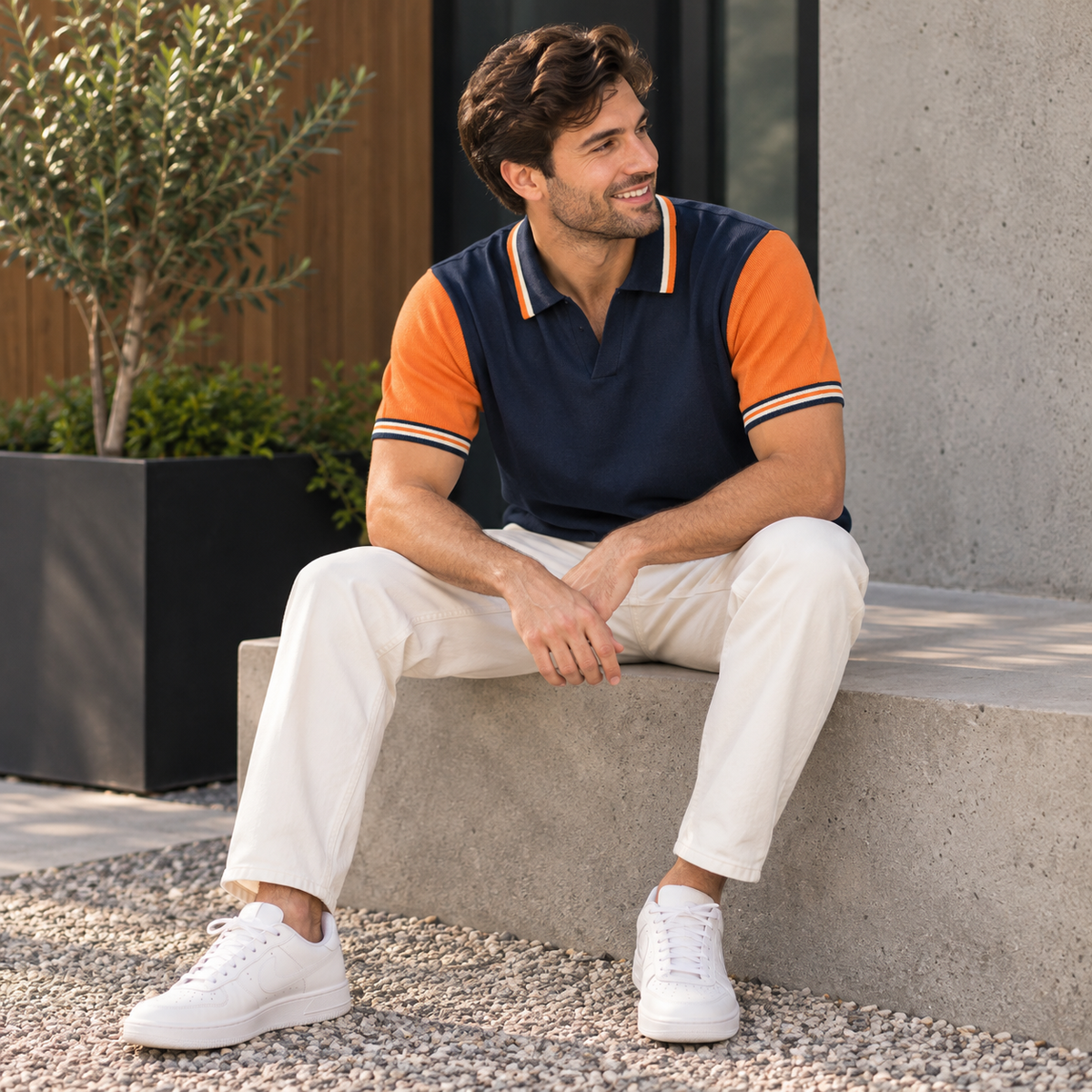 A man sitting on concrete steps outdoors wearing a navy polo shirt with orange sleeves and striped trim, paired with off-white trousers and white sneakers.