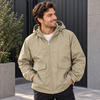 A man wearing a light khaki hooded zip-up jacket with front flap pockets, standing outdoors in front of a dark paneled wall.